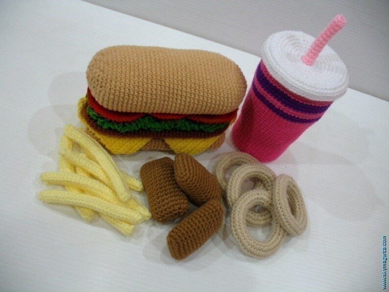 May include: A crocheted hamburger, french fries, onion rings, and a pink and white crocheted soda cup with a pink straw. The food items are all made of yarn and are a fun and whimsical way to represent a classic fast food meal.