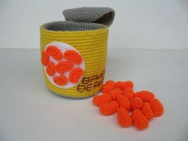 K&ouml;nnte beinhalten: Eine geh&auml;kelte gelbe Dose mit einem grauen Deckel, gef&uuml;llt mit orangefarbenen geh&auml;kelten Bohnen. Die Dose tr&auml;gt den Text "BAKED BEANS" an der Seite.