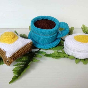May include: Three crocheted zippered pouches shaped like a cup of coffee, a fried egg, and a slice of toast. The coffee cup is blue, the egg is white with a yellow yolk, and the toast is brown with a white egg on top.