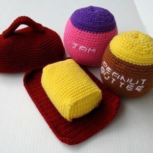 May include: Crocheted plush toys shaped like a loaf of bread, a jar of peanut butter, and a jar of jam. The bread is red, the peanut butter is brown and yellow, and the jam is pink and purple. The toys are labeled "Peanut Butter" and "Jam".