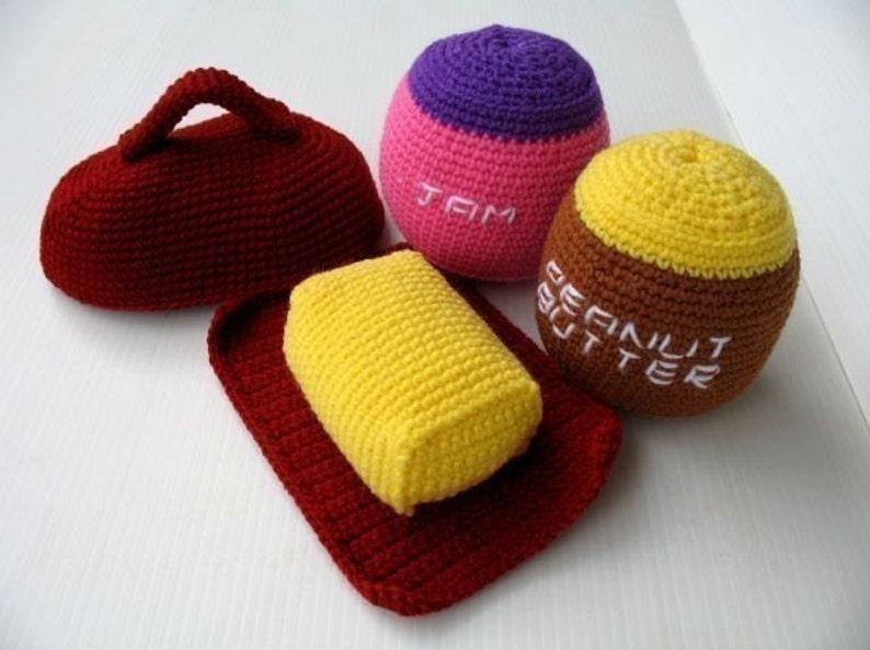 May include: Crocheted plush toys shaped like a loaf of bread, a jar of peanut butter, and a jar of jam. The bread is red, the peanut butter is brown and yellow, and the jam is pink and purple. The toys are labeled "Peanut Butter" and "Jam".