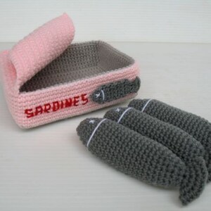 K&ouml;nnte beinhalten: Eine geh&auml;kelte Sardinenb&uuml;chse in Rosa und Grau mit dem Wort "SARDINES" bestickt auf der Vorderseite. Die Dose ist ge&ouml;ffnet und drei graue geh&auml;kelte Sardinen fallen heraus.