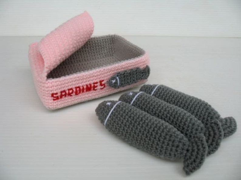 K&ouml;nnte beinhalten: Eine geh&auml;kelte Sardinenb&uuml;chse in Rosa und Grau mit dem Wort "SARDINES" bestickt auf der Vorderseite. Die Dose ist ge&ouml;ffnet und drei graue geh&auml;kelte Sardinen fallen heraus.