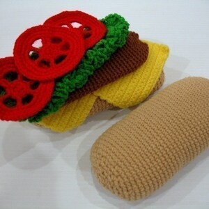 May include: A crocheted sandwich with red tomato slices, green lettuce, brown meat, and yellow cheese. The sandwich is next to a crocheted brown bun.