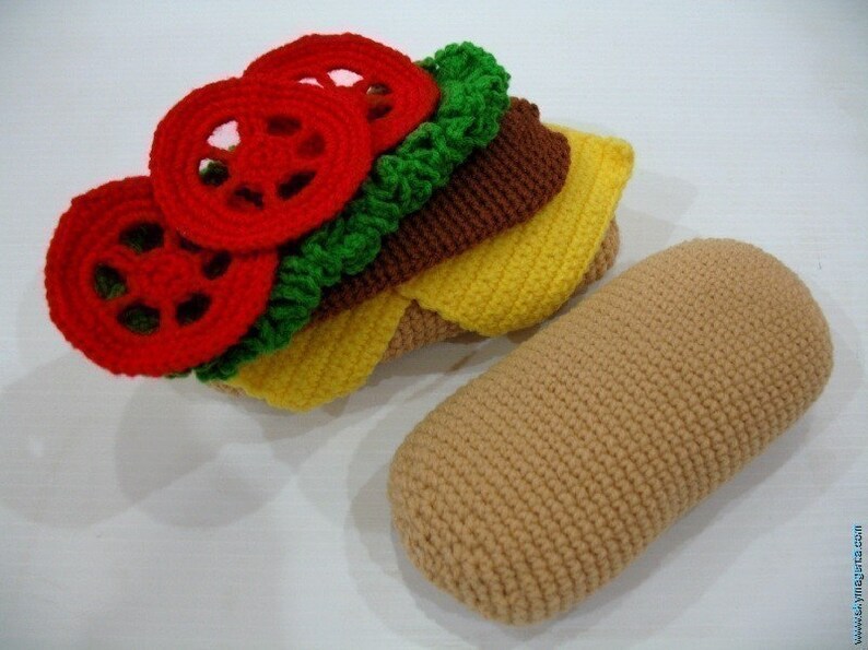 May include: A crocheted sandwich with red tomato slices, green lettuce, brown meat, and yellow cheese. The sandwich is next to a crocheted brown bun.