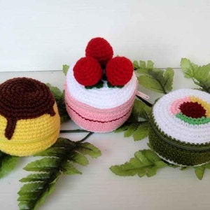 Puede incluir: Tres estuches de cremallera de crochet con temática de comida. Uno es un pudín amarillo y marrón, otro es un pastel rosa y blanco con fresas rojas y el último es un rollo de sushi verde y blanco.