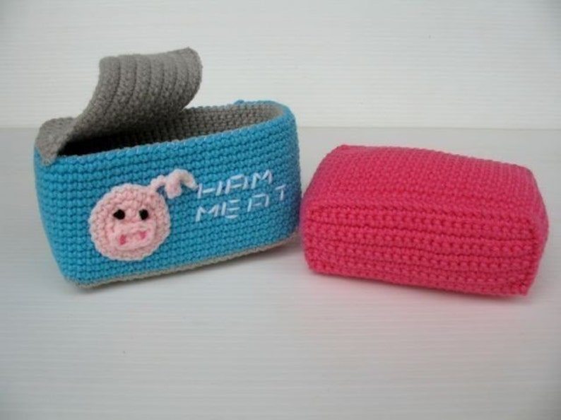 K&ouml;nnte beinhalten: Eine blaue geh&auml;kelte Box mit einem grauen Deckel und einem gestickten rosa Schweinegesicht auf der Vorderseite. Die Box tr&auml;gt die Aufschrift "Liam Meet" in wei&szlig;er Stickerei. Daneben befindet sich eine rosa geh&auml;kelte Box.