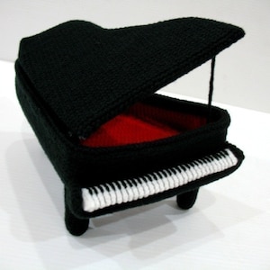 Può includere: Un pianoforte a coda in crochet nero con un interno in feltro rosso e tasti bianchi. Il coperchio è aperto.