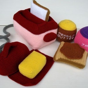 May include: A set of crocheted food items, including a toaster, bread, peanut butter, and jam. The toaster is pink and red with a white bread slice inside. The bread is brown with a yellow butter slice and a brown peanut butter spread. The peanut butter jar is yellow and brown with the text "Peanut Butter". The jam jar is pink and purple with the text "Jam".