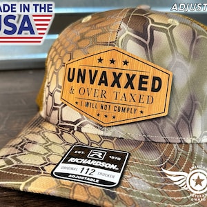 Puede incluir: Gorra de camionero de camuflaje con un parche de madera que dice "UNVAXXED & OVER TAXED I WILL NOT COMPLY". La gorra tiene una parte trasera de malla y una correa ajustable. Hecho en los EE. UU.
