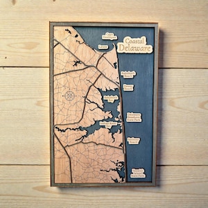 May include: Wooden map of Coastal Delaware, featuring laser-cut details of the coastline, roads, and place names. The map is framed in a dark gray border and includes the text "Coastal Delaware". The map includes the names of the following locations: Cape Henlopen, Lewes, Rehoboth Beach, Dewey Beach, Indian River Bay, Delaware Seashore State Park, Bethany Beach, and Fenwick Island State Park.