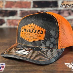 Może przedstawiać: Czarno-pomarańczowa czapka trucker z kamuflażem i regulowanym paskiem. Czapka ma brązową skórzaną naszywkę z tekstem "UNVAXXED & OVER TAXED I WILL NOT COMPLY".