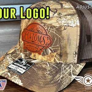 May include: Camouflage trucker hat with a brown mesh back and a leather patch. The patch features the text "CUSTOMS ENGRAVING" and the words "YOUR LOGO!" are displayed above the hat. The hat is adjustable.