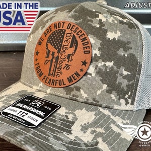 Peut inclure: Un chapeau de camionneur camouflage avec un patch en cuir marron qui dit "We are not descended from fearful men" avec un crâne et des os croisés et un drapeau américain. Le chapeau est réglable et a un dos en maille.