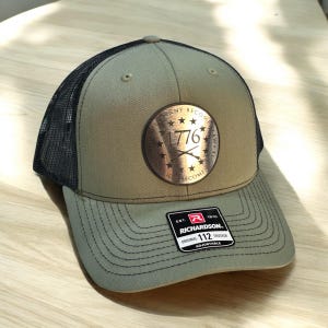 Peut inclure: Casquette de camionneur vert olive et noir avec un écusson rond en bronze portant l'inscription "1776" et des fusils croisés. La casquette a une visière incurvée et un dos en filet. L'étiquette Richardson est visible.
