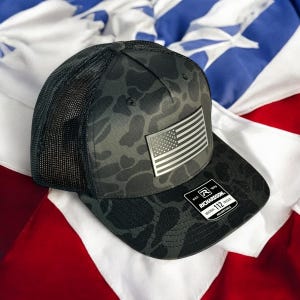 May include: A black and gray camouflage trucker hat with a mesh back. The hat features a silver patch of the American flag and a Richardson 112 tag. The hat is displayed on a red, white, and blue background.