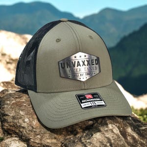 Peut inclure: Casquette de camionneur vert olive avec un dos en filet noir. La casquette présente un emblème argenté avec le texte "UNVAXXED & OVER TAXED I WILL NOT COMPLY." Une étiquette Richardson 115 Trucker est visible.