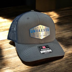 Peut inclure: Casquette de camionneur grise avec un dos en filet bleu marine. La casquette porte l'inscription "UNVAXXED & OVER TAXED I WILL NOT COMPLY" dans un design argenté et bleu. La casquette est réglable.