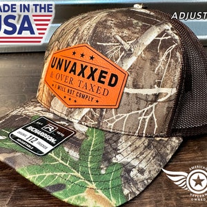 May include: A camouflage trucker hat with a brown mesh back. The hat has a brown leather patch with the text "UNVAXXED & OVER TAXED I WILL NOT COMPLY" in black lettering. The hat is adjustable.