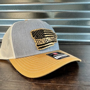 以下が含まれることがあります： グレーとタンのトラッカーハット。ゴールドメタルのパッチには、使い古したアメリカの旗と「We the People」の文字が描かれています。