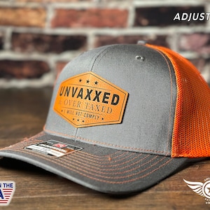 Może przedstawiać: Szara i pomarańczowa czapka trucker z brązową skórzaną naszywką z napisem "Unvaxxed & Over Taxed I Will Not Comply". Czapka jest regulowana.