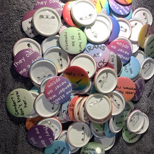 Puede incluir: Un conjunto de botones redondos y coloridos con varios textos y diseños. Los botones presentan frases como "They call me a demi god", "I ain't here to be romantic" y "Gender was never an option". Los pines son en tonos morado, azul, verde y rojo.