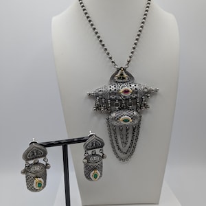 May include: A silver-toned necklace and earring set displayed on a mannequin. The necklace features a pendant with red, green, and black gemstones. The earrings are a matching design. The jewelry has intricate detailing and a vintage aesthetic.