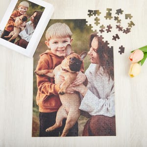 May include: A jigsaw puzzle featuring a photo of two people and a dog. The people are a young boy and a woman. The boy is wearing a brown sweater and the woman is wearing a white sweater. The dog is a white and brown French Bulldog. The puzzle is on a wooden surface.