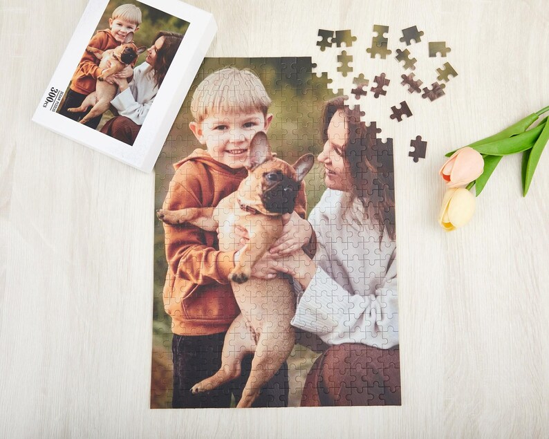 May include: A jigsaw puzzle featuring a photo of two people and a dog. The people are a young boy and a woman. The boy is wearing a brown sweater and the woman is wearing a white sweater. The dog is a white and brown French Bulldog. The puzzle is on a wooden surface.