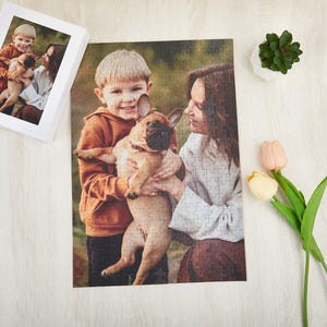 May include: A family of three, a woman and two children, are smiling and holding a small, light brown and white dog. The dog is sitting on the woman's lap and the children are holding the dog's paws. The family is standing in a grassy field.