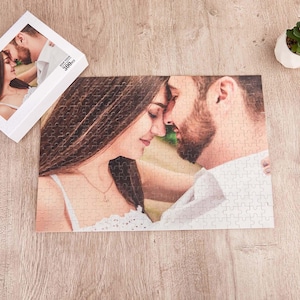 Könnte beinhalten: Ein rechteckiges Puzzle mit einem Fotodruck eines sich umarmenden Paares, wobei die Frau ein weißes Oberteil und der Mann ein weißes Hemd trägt. Die Puzzleschachtel ist in der Ecke sichtbar, mit 300PCS beschriftet und zeigt dasselbe Bild.