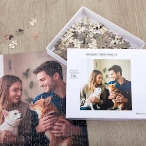 May include: A 196-piece wooden jigsaw puzzle featuring a photograph of a person with long blonde hair and a person with short dark hair, each holding a small dog. The white box displays the text HIGH QUALITY WOOD PRODUCTS, JIGSAW PUZZLES, 196, and SIZE: 280x280mm.
