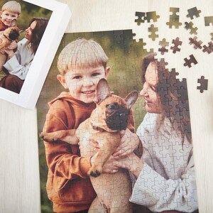 May include: A photo puzzle featuring a family with a brown and white French bulldog. The puzzle shows a young boy holding the dog, a woman standing behind him, and a smaller image of a young boy holding the dog in a frame.