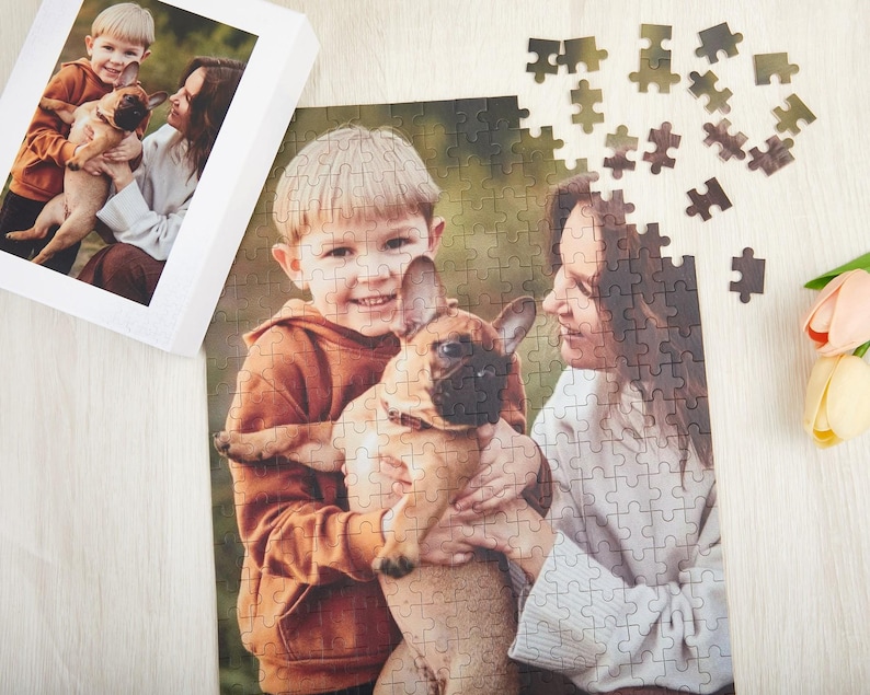 May include: A photo puzzle featuring a family with a brown and white French bulldog. The puzzle shows a young boy holding the dog, a woman standing behind him, and a smaller image of a young boy holding the dog in a frame.