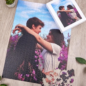 Könnte beinhalten: Ein rechteckiges Puzzle mit einem Foto von zwei Personen, die sich in einem Feld aus violetten Blumen unter einem blauen Himmel umarmen. Das Puzzle ist teilweise zusammengesetzt, mit einigen losen Teilen und einer weißen Schachtel mit dem Bild.