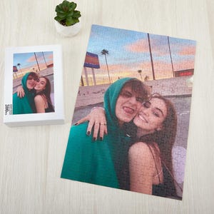 May include: A couple pose for a photo in front of a sunset backdrop. The man is wearing a green hoodie and the woman is wearing a white vest top. The photo is printed on a jigsaw puzzle.