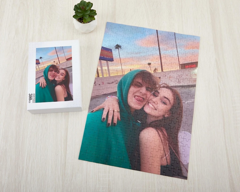 May include: A couple pose for a photo in front of a sunset backdrop. The man is wearing a green hoodie and the woman is wearing a white vest top. The photo is printed on a jigsaw puzzle.
