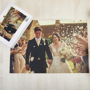 Könnte beinhalten: Ein 300-teiliges Puzzle mit einem Hochzeitsfoto eines lächelnden Paares, das durch Konfetti geht. Das Puzzle liegt neben seiner Schachtel, die ebenfalls das Bild zeigt. Das Paar ist von Gästen umgeben. Eine kleine Pflanze und Blumen sind im Hintergrund.
