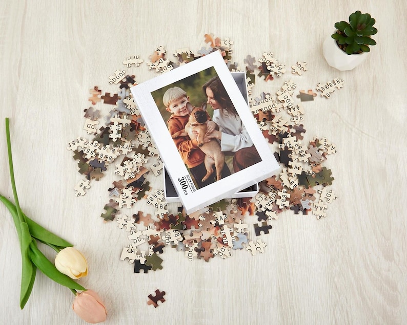May include: A 300 piece jigsaw puzzle featuring a photo of a woman and a child with a dog. The puzzle box is white with black text and a photo of the puzzle image.
