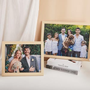 May include: Two framed jigsaw puzzles featuring family photographs. One depicts a wedding couple, the other a group portrait. Both puzzles are in light wood frames, displayed on a white surface with a small plant and books.