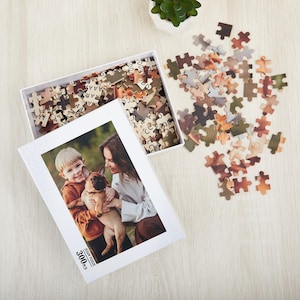 May include: A white box with a 3000 piece jigsaw puzzle inside. The puzzle features a photo of two people and a dog. The box is open and the puzzle pieces are scattered around it.