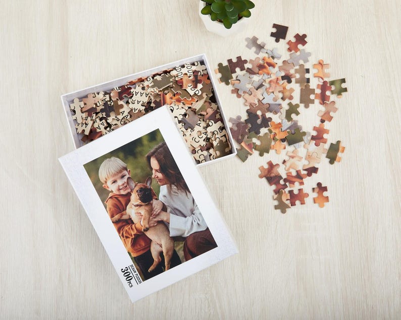 May include: A white box with a 3000 piece jigsaw puzzle inside. The puzzle features a photo of two people and a dog. The box is open and the puzzle pieces are scattered around it.