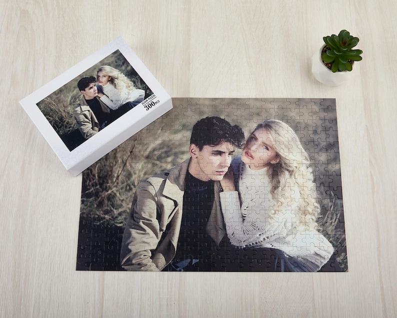 May include: A 300-piece jigsaw puzzle featuring a couple posing together in a woodland setting. The puzzle is made of cardboard and has a matt finish.