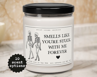 May include: White candle jar with a black lid and a label that reads "Smells like you're stuck with me forever". The label features a black and white illustration of two skeletons holding hands. The candle is made with all natural soy wax and is hand-poured in the USA.