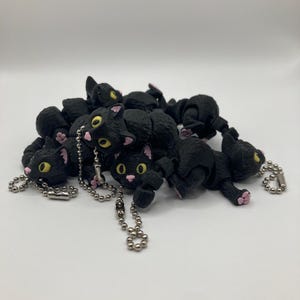 May include: A group of black cat keychains with yellow eyes and pink paws. The keychains are made of plastic and have a silver chain.