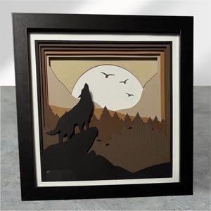 May include: A framed, layered paper art piece featuring a howling wolf silhouette on a rock, set against a sunset with mountains and trees. The artwork uses shades of brown, tan, and black, with a white border and a black frame.