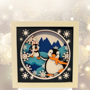 May include: A 3D papercraft scene with two penguins in a winter landscape. The penguins are wearing scarves and hats. The scene is framed in a white frame and features a blue and white color scheme.