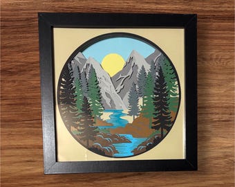 Könnte beinhalten: Gerahmtes Papierkunstwerk mit einer Berglandschaft. Das kreisförmige Kunstwerk zeigt einen Fluss, Bäume, Berge und eine Sonne. Der Rahmen ist schwarz und der Hintergrund ist hellbeige. Ein dekoratives Kunstwerk.