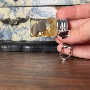 May include: A small clear glass vial keychain filled with clear resin, containing two smooth grey stones and several dried yellow flowers. The vial is sealed with a silver-colored metal screw cap and features a silver-toned metal lobster clasp attachment.