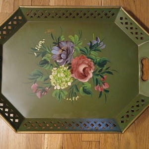 May include: An octagonal olive green metal serving tray featuring a hand-painted floral bouquet in shades of purple, pink, and white. The tray has a lattice-style cutout border, gold-toned edges, and two integrated side handles for carrying.